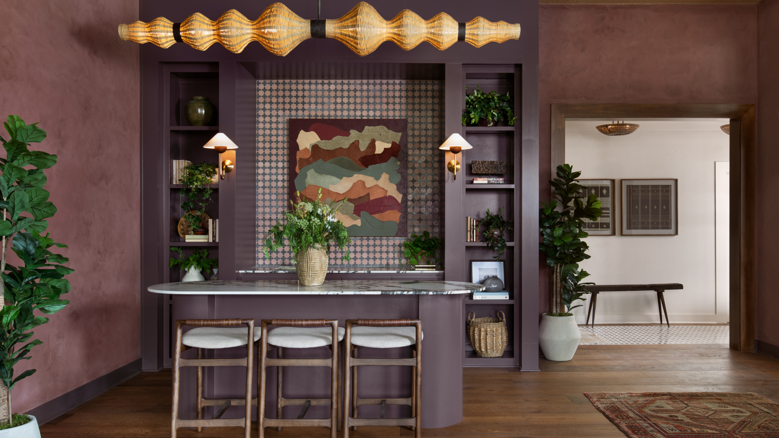 A stylish kitchen with a purple island, three stools, gold pendant light, abstract wall art, built-in shelves, and potted plants in a modern, earthy setting.