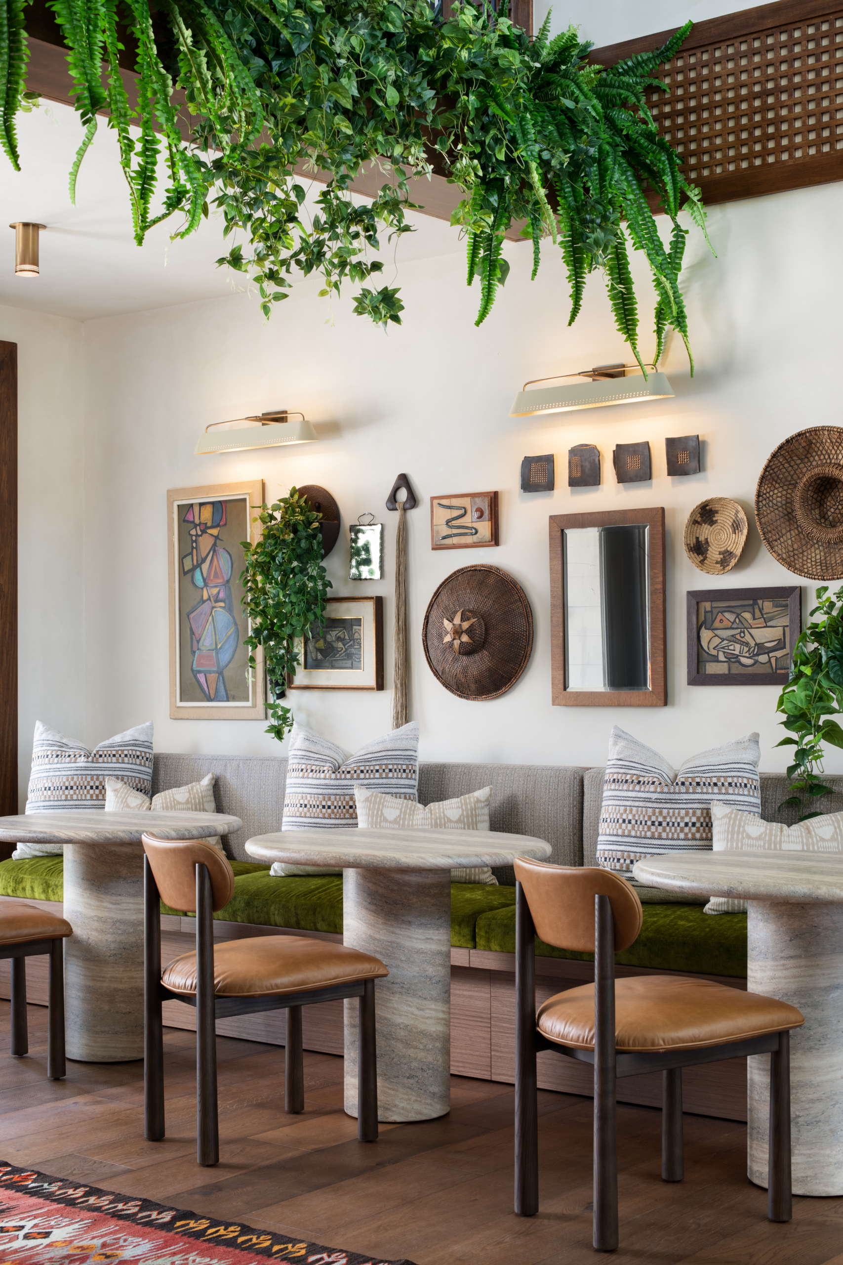 A cozy cafe with wooden tables and chairs, green cushioned bench seating, wall art, woven baskets, and lush hanging plants, creating a warm, inviting atmosphere.