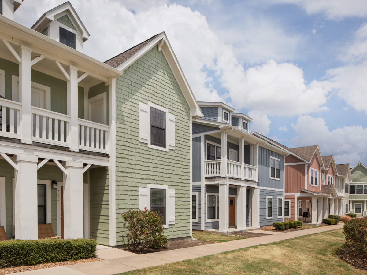 A row of colorful, modern two-story houses with front porches and balconies on a sunny day, with a sidewalk and bushes in front.