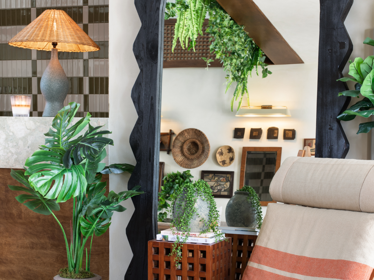 A cozy room with a striped lounge chair, large black-framed mirror, potted plants, wall art, and a lamp with a woven shade. Ceiling plants hang near geometric tiles, creating a modern, natural atmosphere.