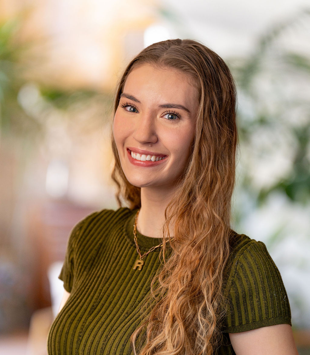 A young woman with long, wavy blonde hair smiles at the camera. She is wearing a ribbed olive-green top and a necklace, standing indoors with a blurred background and green plants.