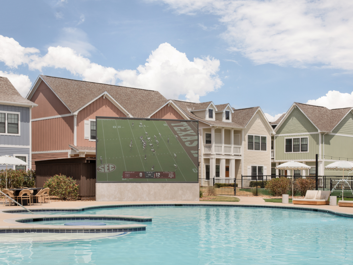 A poolside area with lounge chairs and umbrellas, featuring a large outdoor screen showing a football game. Colorful houses are in the background under a partly cloudy sky.