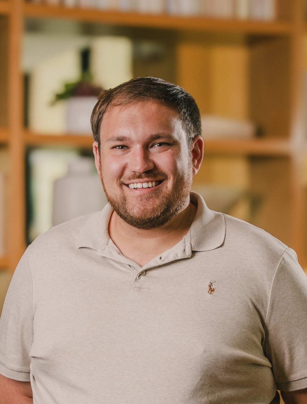 A man with short brown hair and a beard smiles at the camera, wearing a light beige polo shirt. He stands indoors in front of shelves filled with books and decor.