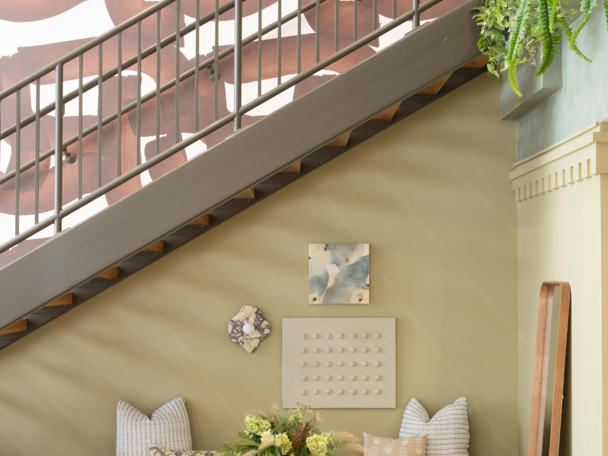 A cozy seating area under a staircase features two wooden chairs, a low coffee table with flowers, and a bench with throw pillows against a sage green wall, showcased in the before and after rambler college park south transformation.