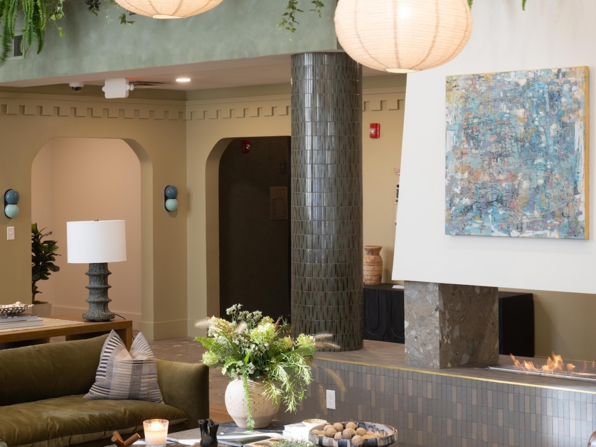 A modern lounge area with a fireplace, abstract art, globe pendant lights, and greenery hanging from a mezzanine railing above, part of the before and after Rambler College Park South renovation.
