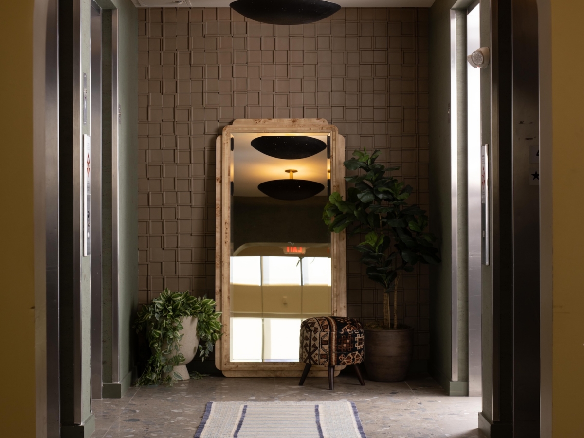 An elevator lobby at Rambler College Park South featuring a long runner rug leading to a floor mirror, a small patterned stool, and potted plants centered against a textured wall.
