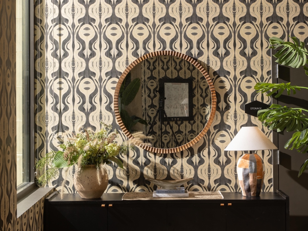 A black console table sits against a patterned wallpapered wall, a before and after detail of the College Park South rambler, featuring a round mirror, a vase of flowers, a table lamp, and a stack of books.