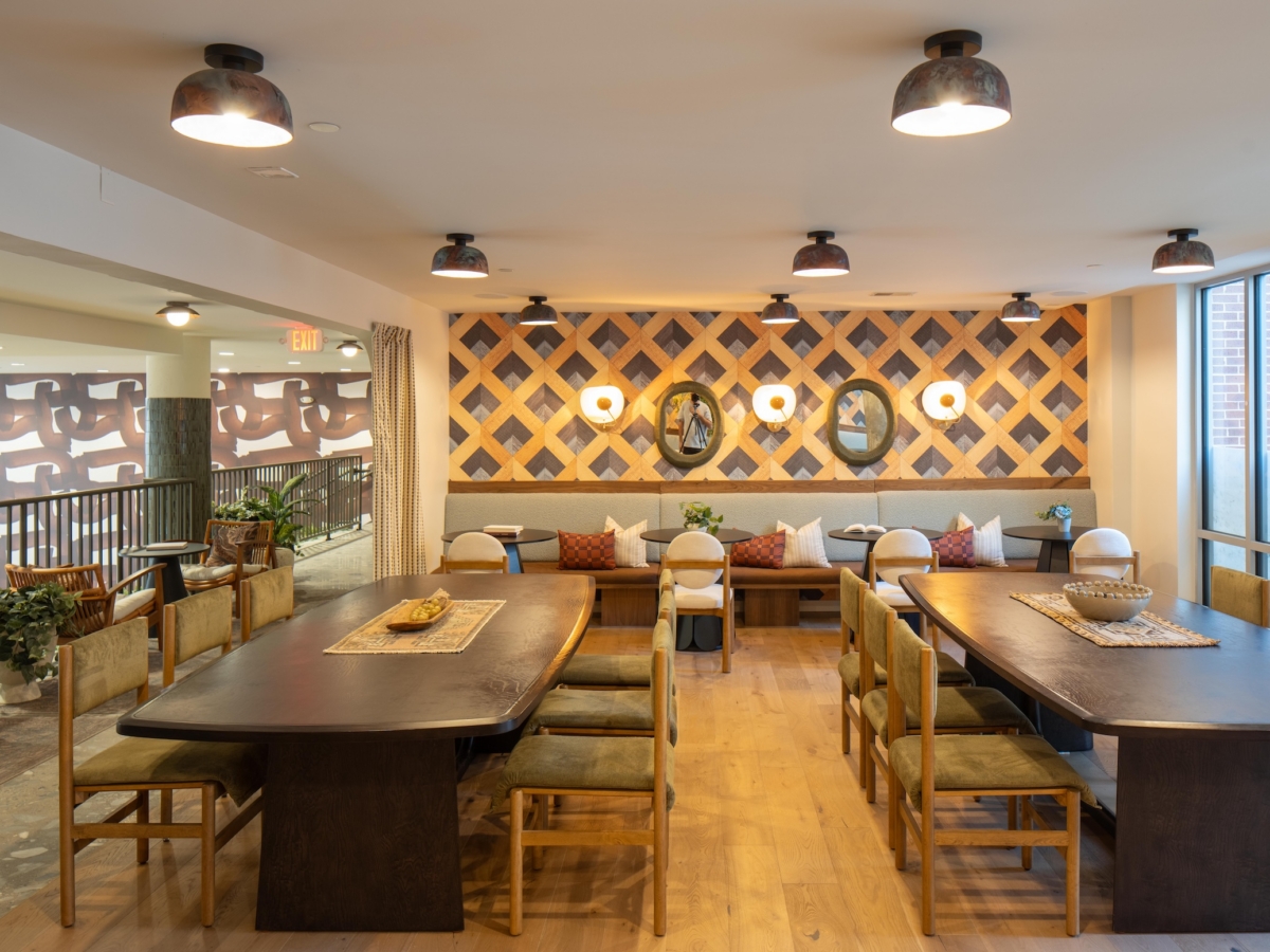 A lounge area with two large wooden tables, green upholstered chairs, and a long bench seat against a wall with geometric patterns and mirrors, captured in a before and after at Rambler College Park South.