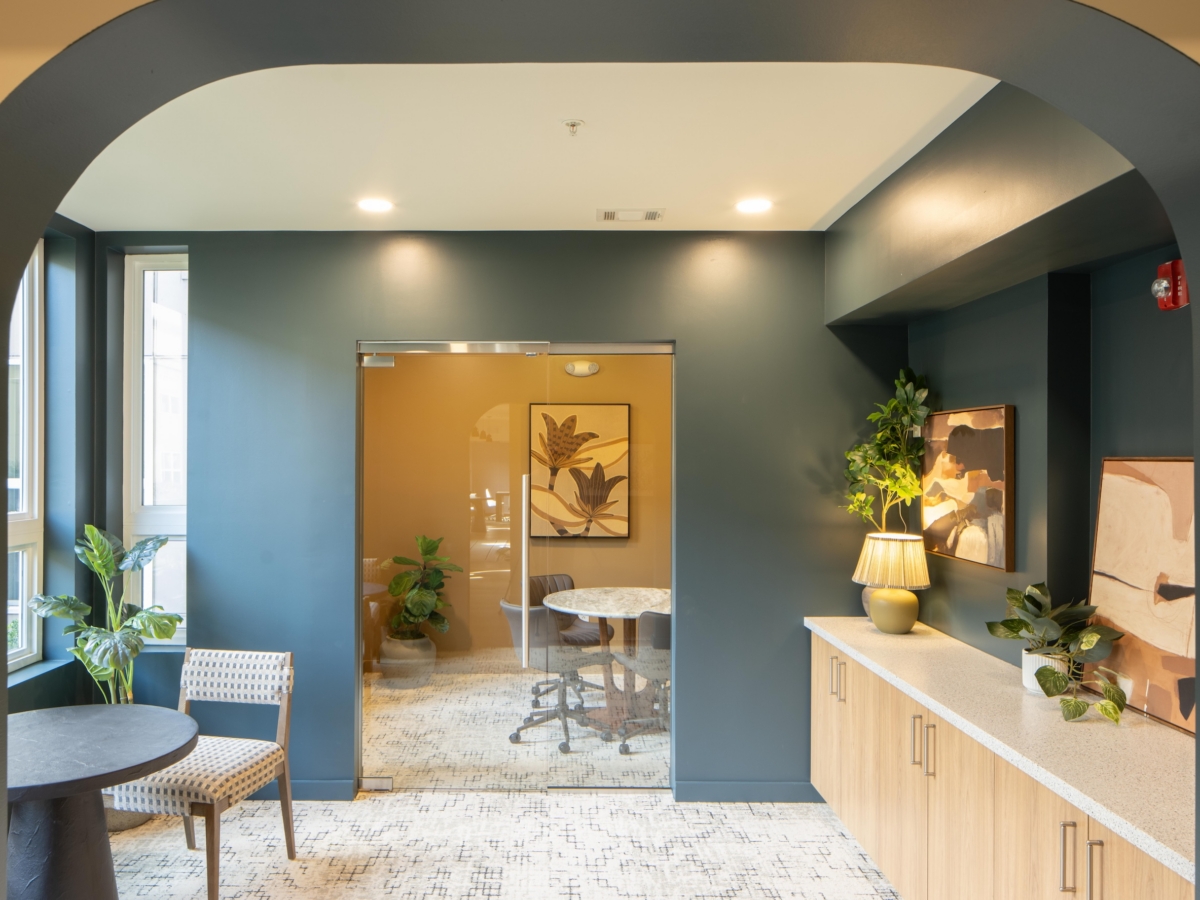 A modern office lounge at Rambler College Park South featuring dark blue walls, a glass-walled conference room, and a wooden built-in cabinet with decorative lamps and artwork.