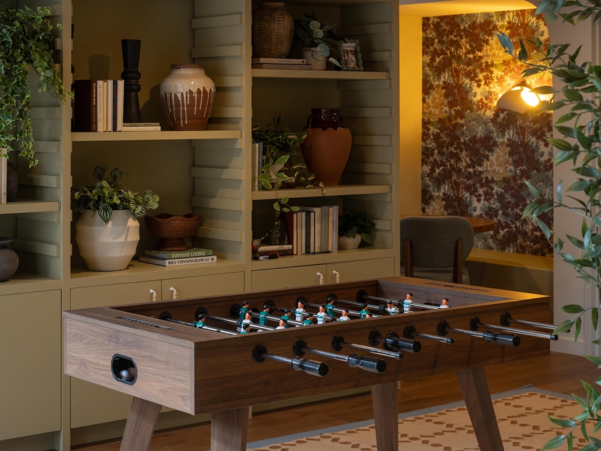 A wooden foosball table sits on a patterned rug in front of olive green shelves, showcasing a before and after transformation in this College Park South rambler.
