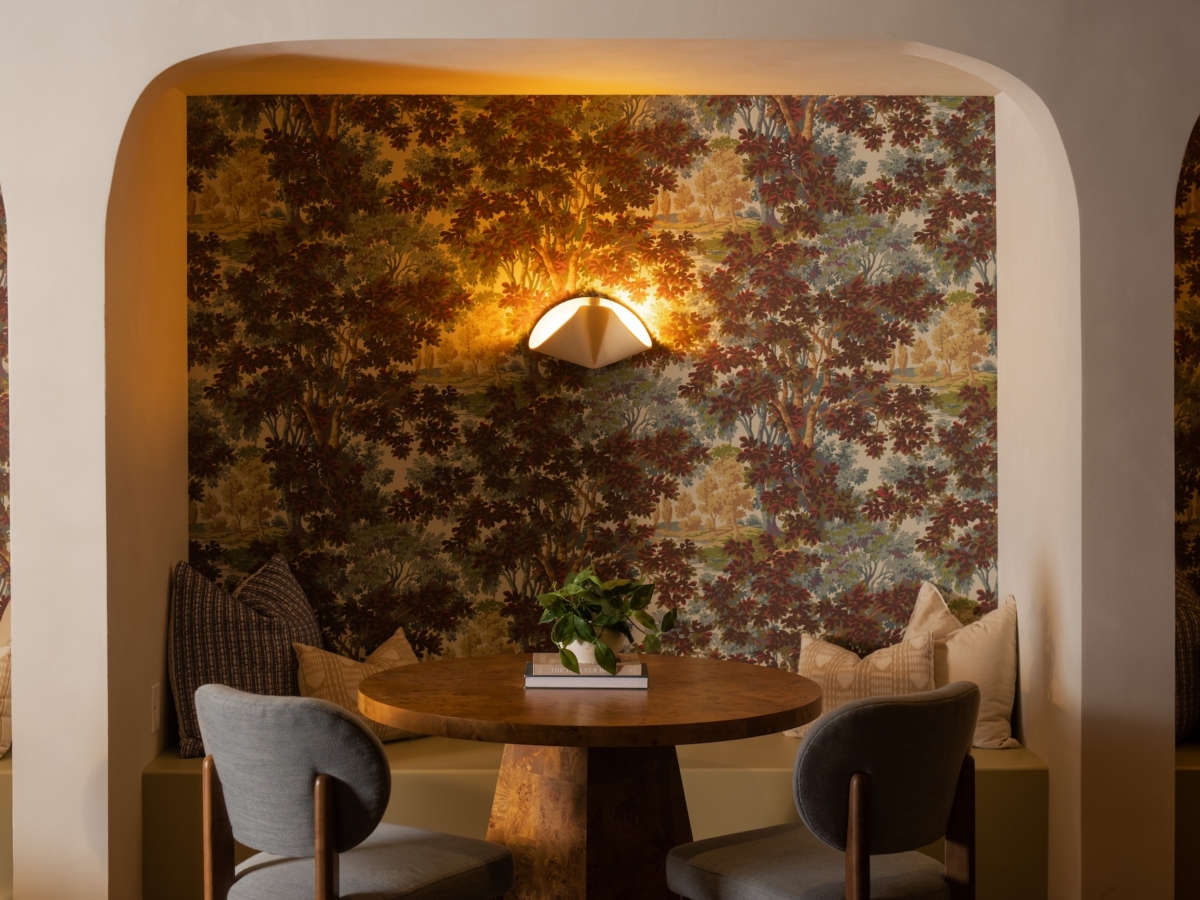 A circular wooden table with a small potted plant and books sits in a recessed nook lined with forest-patterned wallpaper, two grey chairs, and a wall-mounted light fixture, showcasing the before and after of this College Park South rambler.